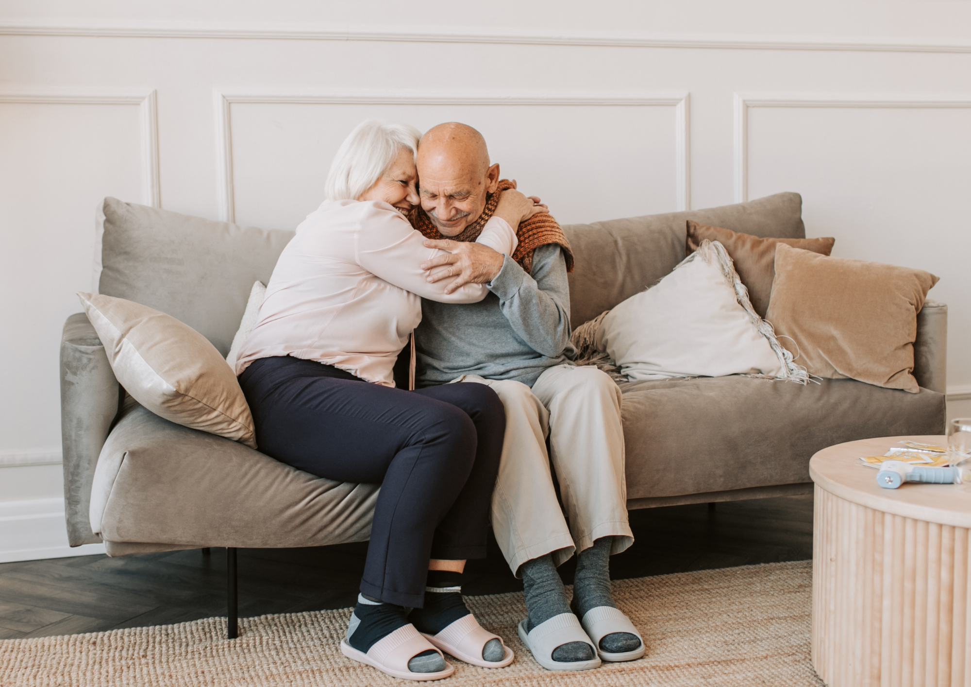 An elderly woman warmly hugging an older man as they sit together on a cozy living room sofa.