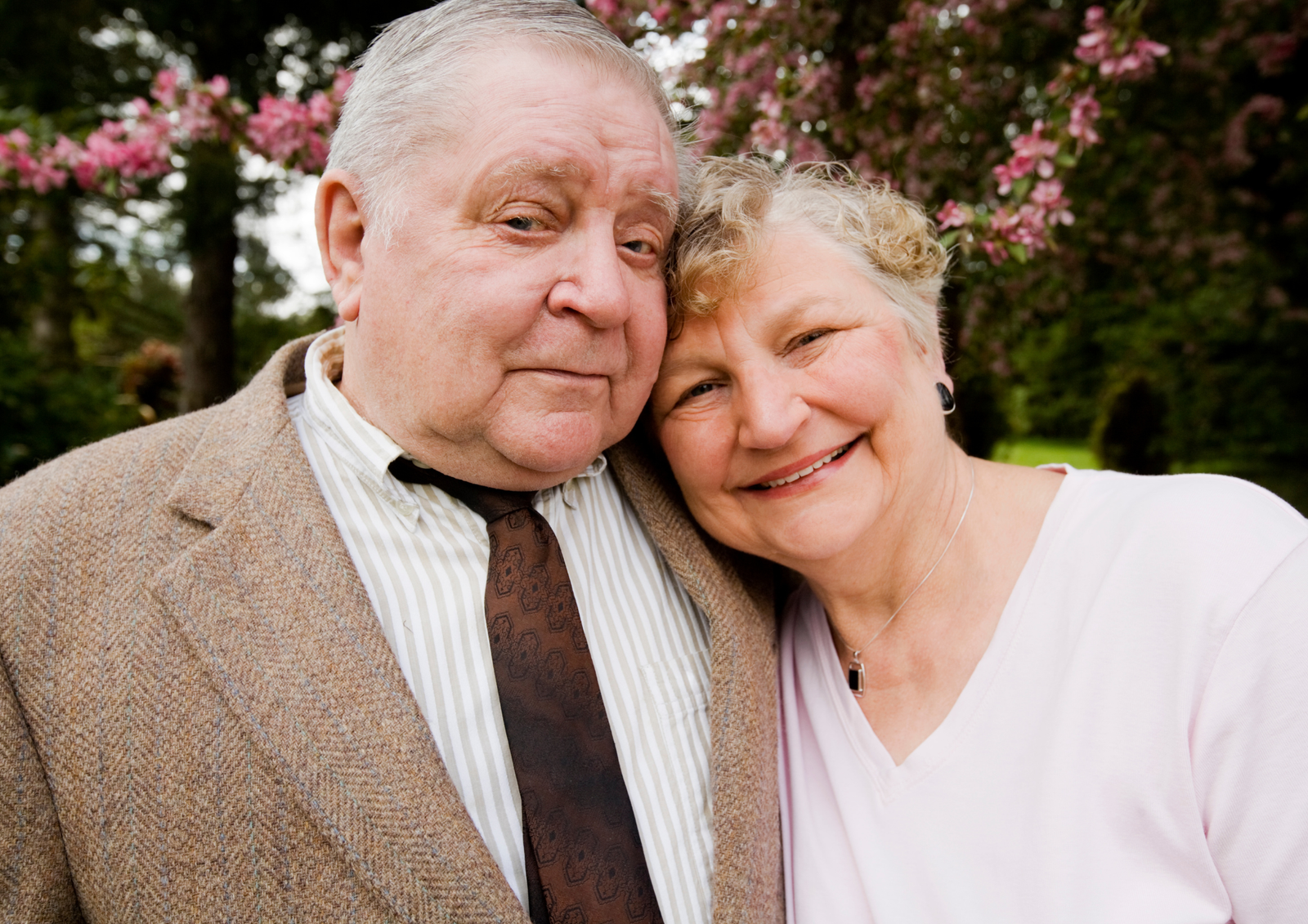 Senior couple smiling together outdoors in front of a blossoming pink tree.