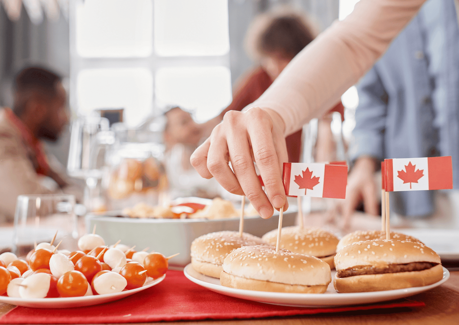 Canada Day Gift Baskets: A hand placing a tiny Canadian flag toothpick into sliders on a party table with appetizers.