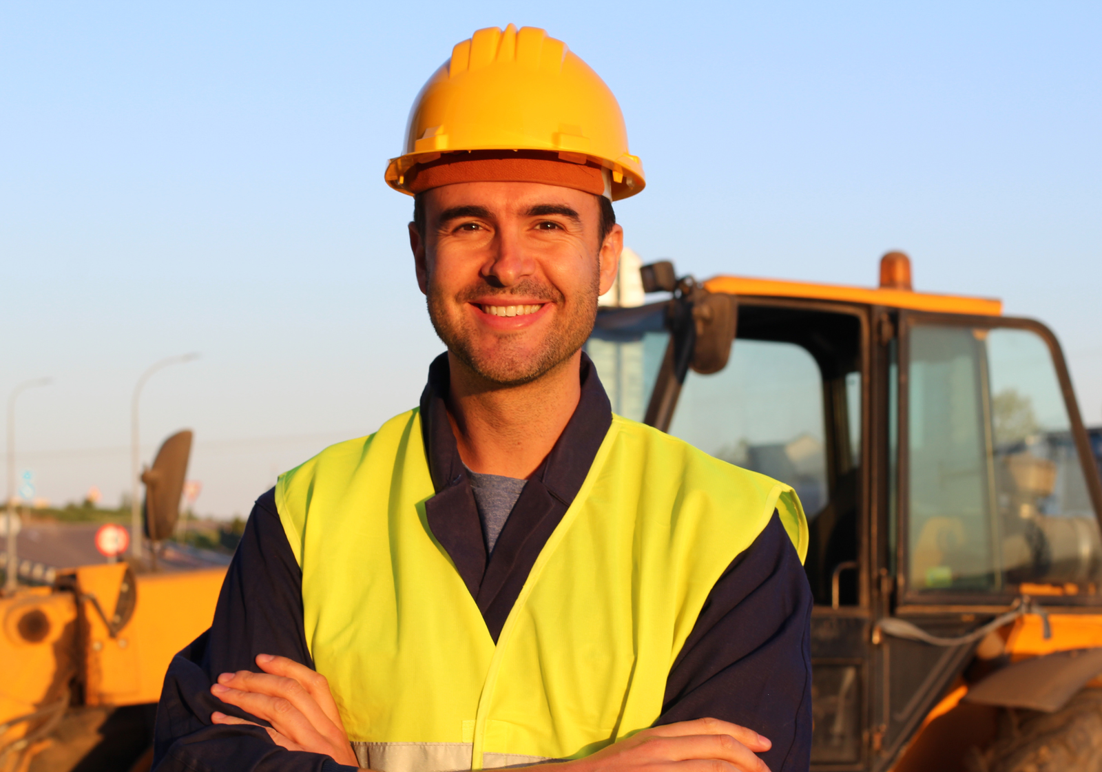 Smiling male construction worker wearing a hardhat and high-visibility vest standing in front of heavy machinery.