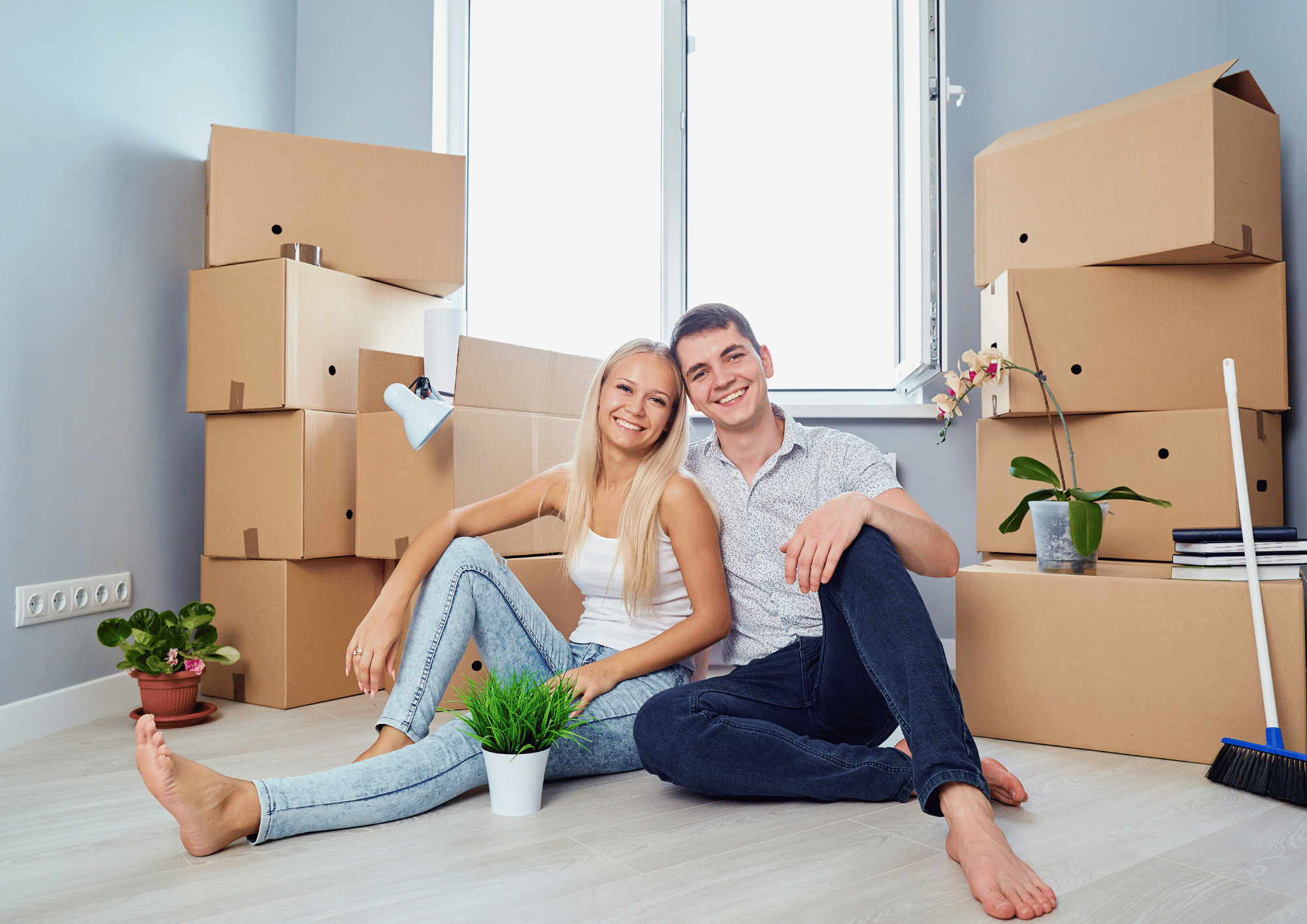 A happy young couple sitting on the floor of their new home surrounded by moving boxes and potted plants.