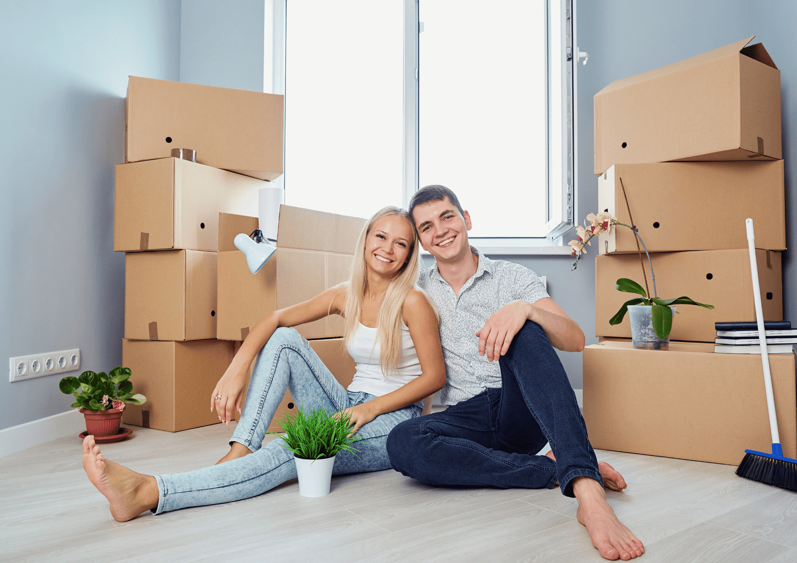 A happy young couple sitting on the floor of their new home surrounded by moving boxes and potted plants.