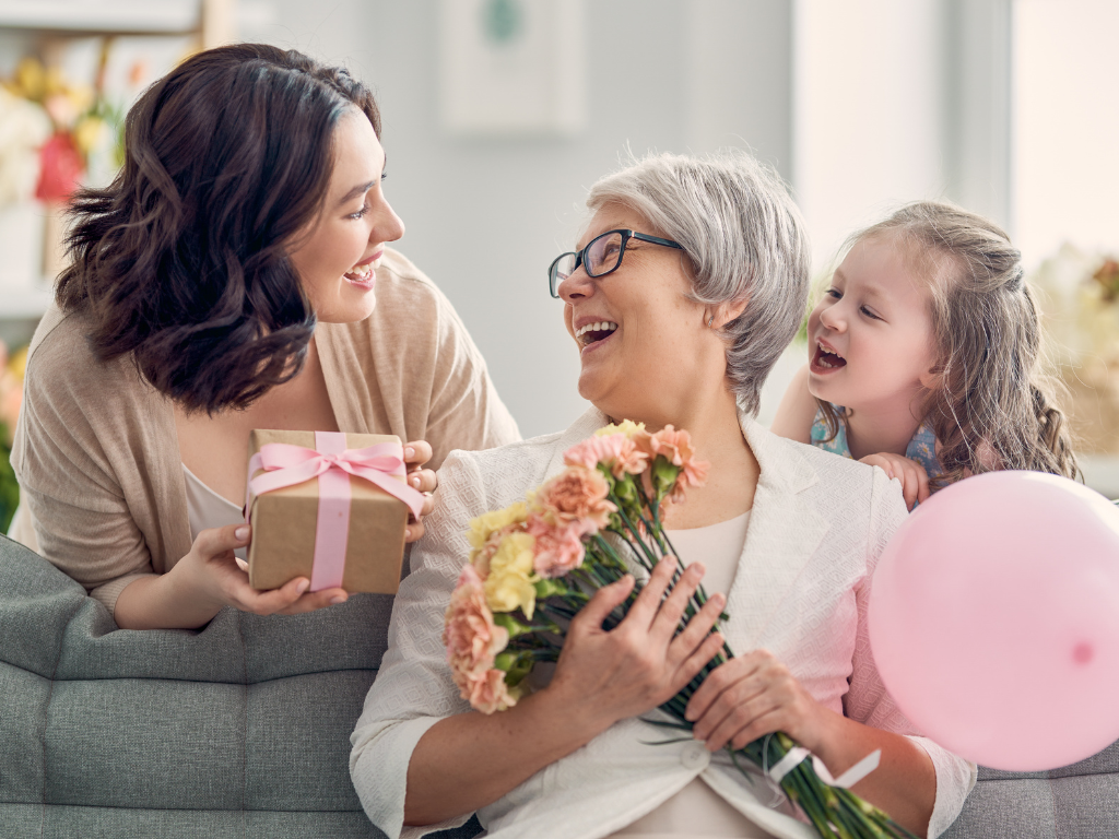 Three generations of women laughing together as a daughter gives a wrapped gift and a granddaughter holds a pink balloon for Mother's Day.