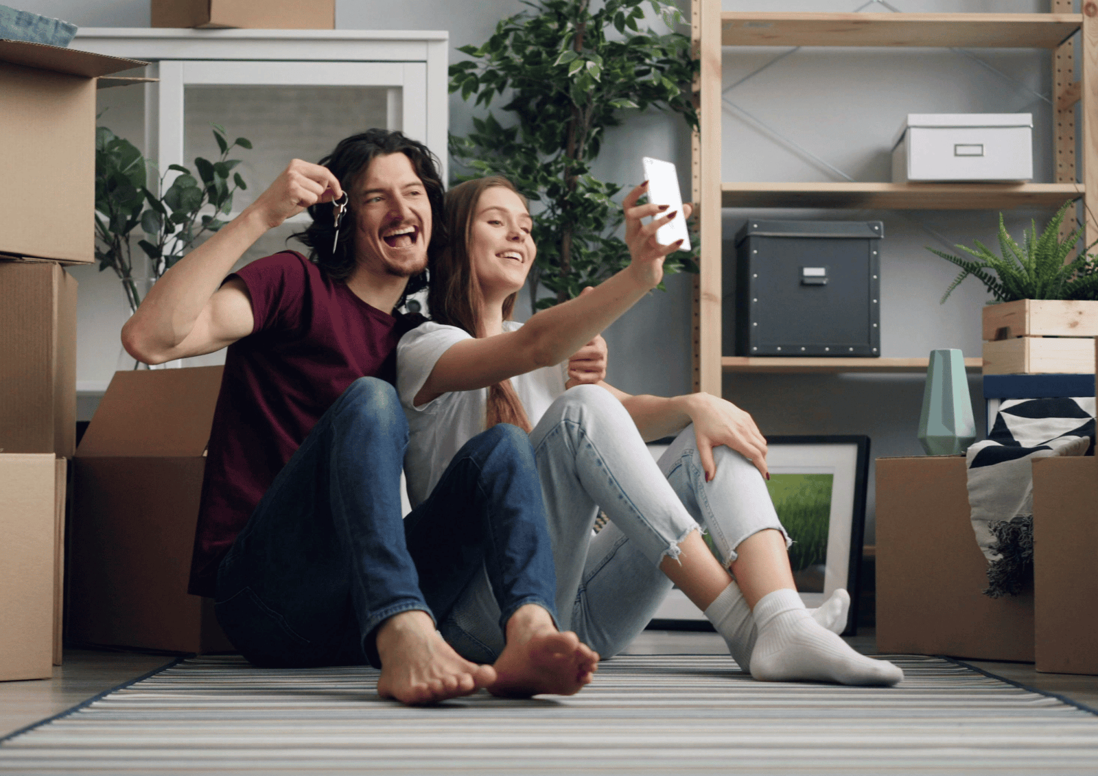 A happy couple sitting on the floor of their new home taking a selfie while holding up a pair of house keys.