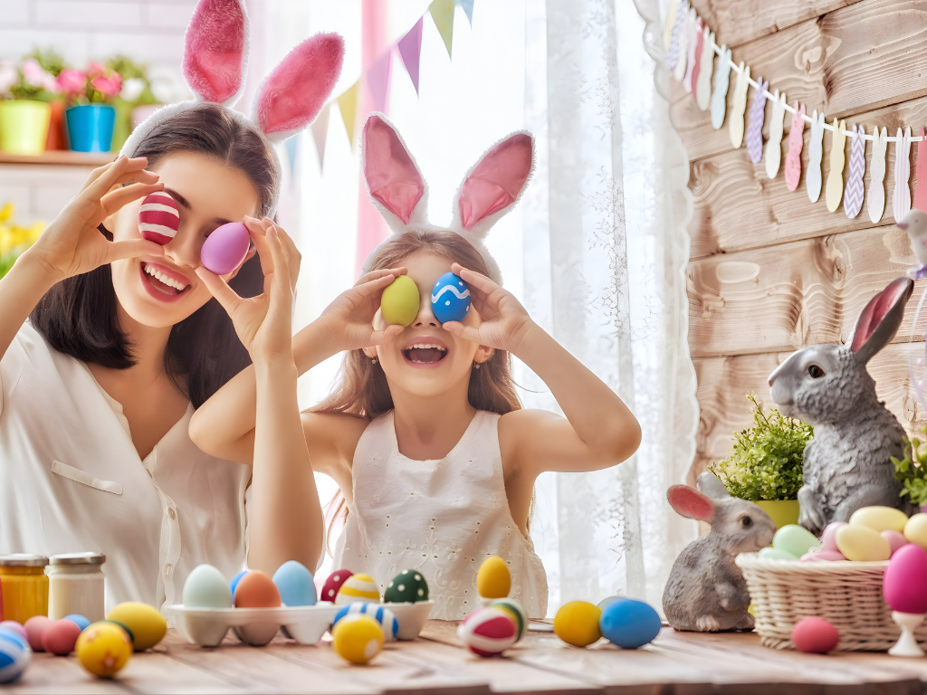 A mother and daughter wearing white bunny ears and laughing while holding colorful painted Easter eggs over their eyes.