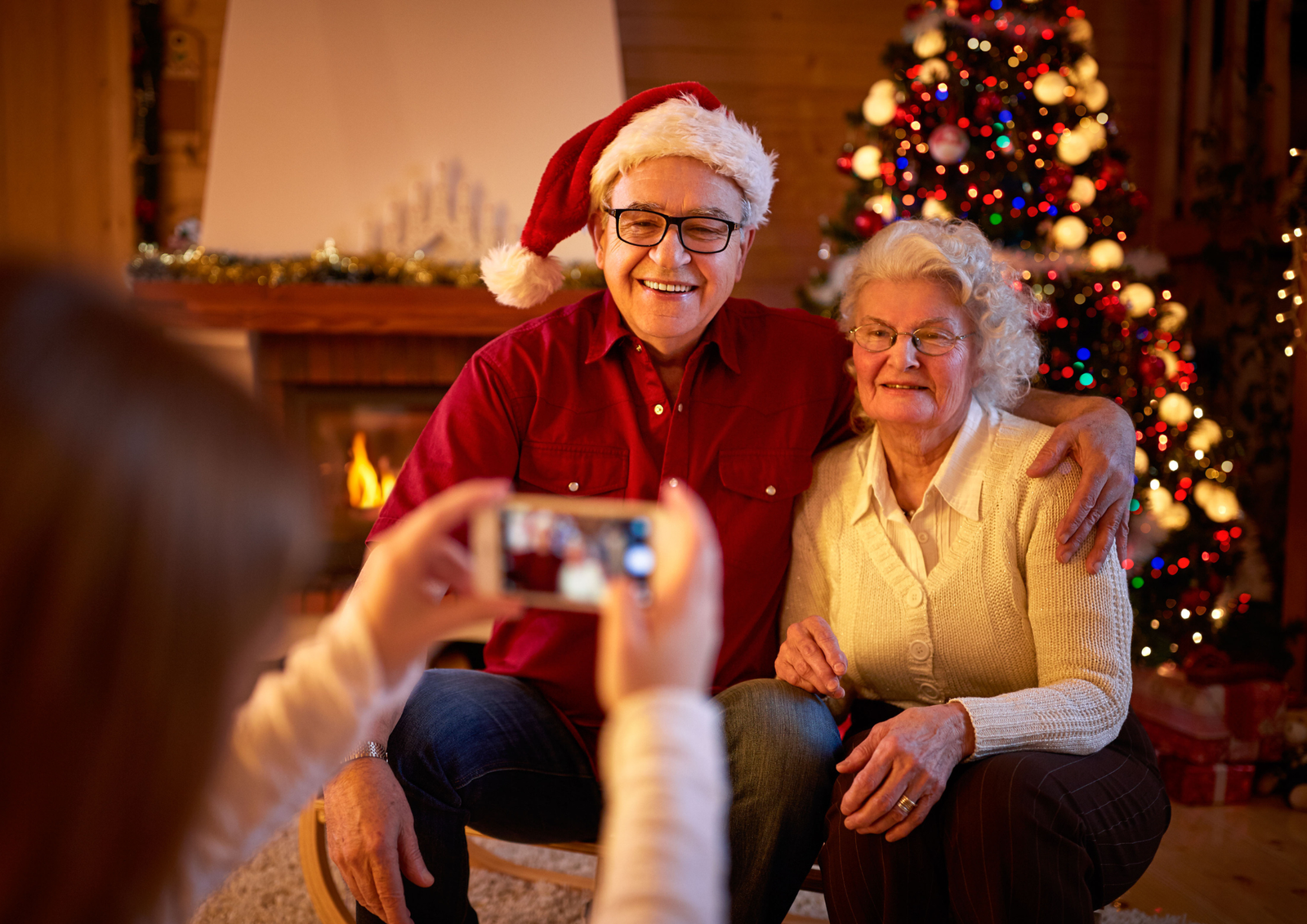 Smiling elderly couple wearing a Santa hat near a lit fireplace, illustrating first Christmas gift ideas for in-laws.