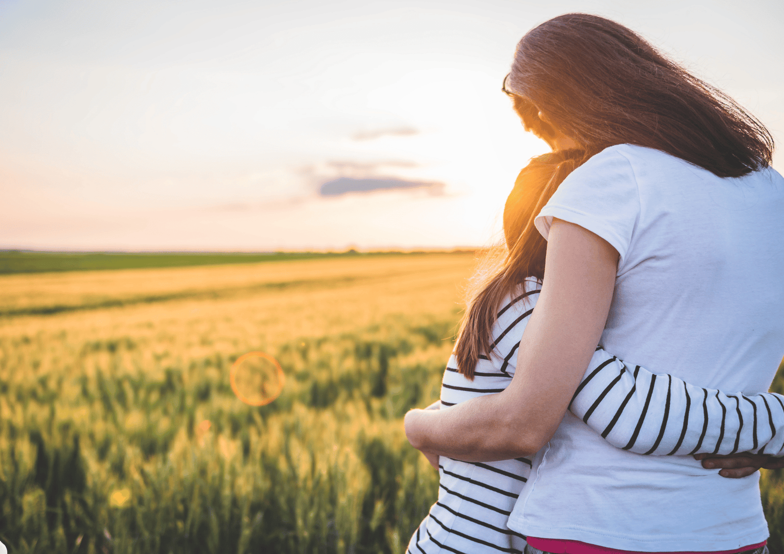 A woman and young girl hugging in a golden wheat field at sunset, representing comfort and sympathy.