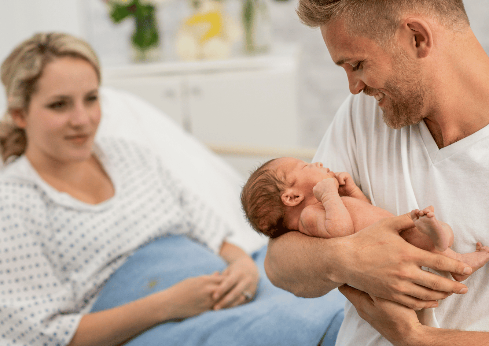 Best Gift Baskets for New Dads: A smiling father cradling his newborn baby in a hospital room beside his partner.