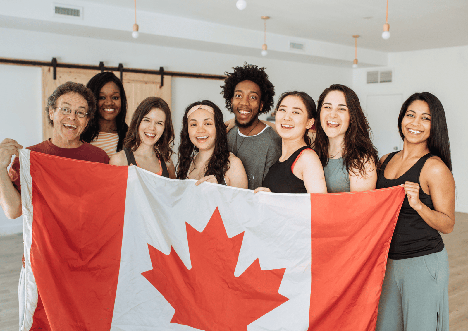 Canada Day Host Gifts: A diverse group of friends smiling and holding a large Canadian flag together indoors.