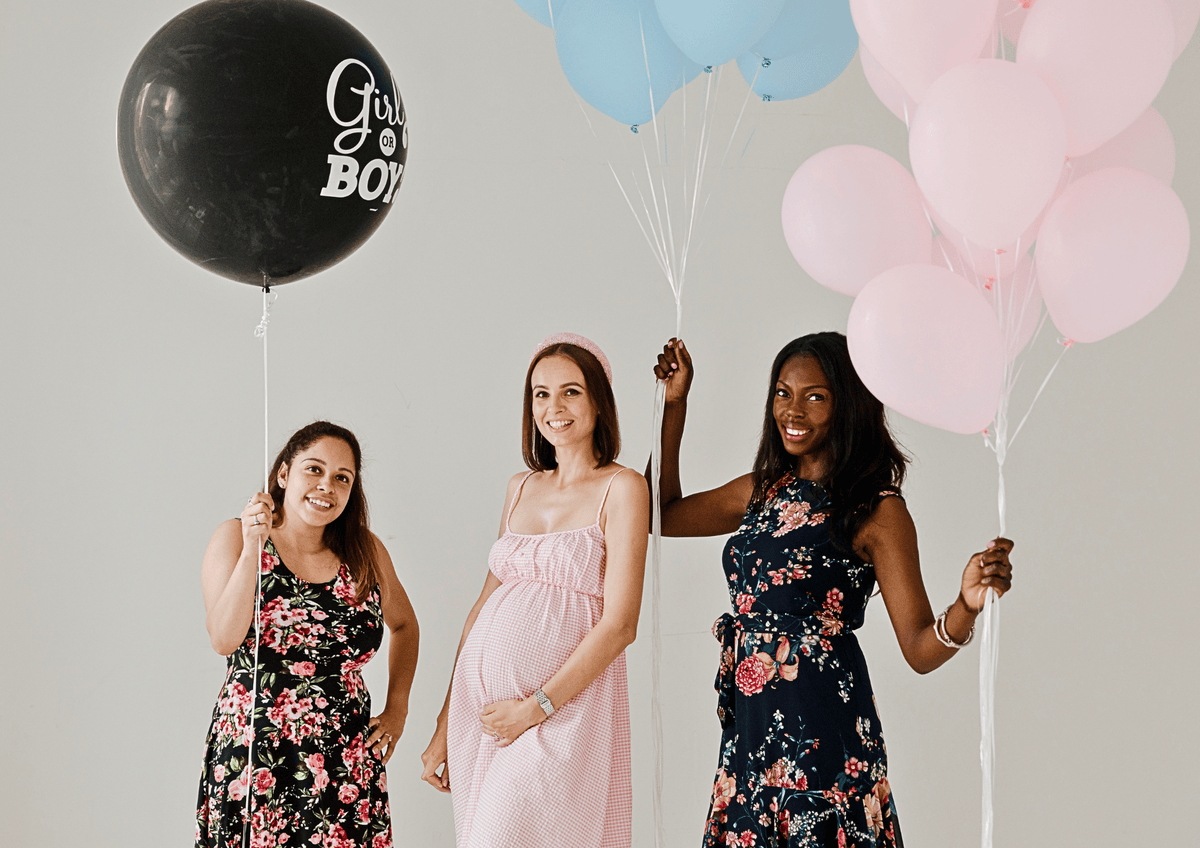 Three smiling women holding pink, blue, and a large black "Girl or Boy" reveal balloon at a baby shower party.