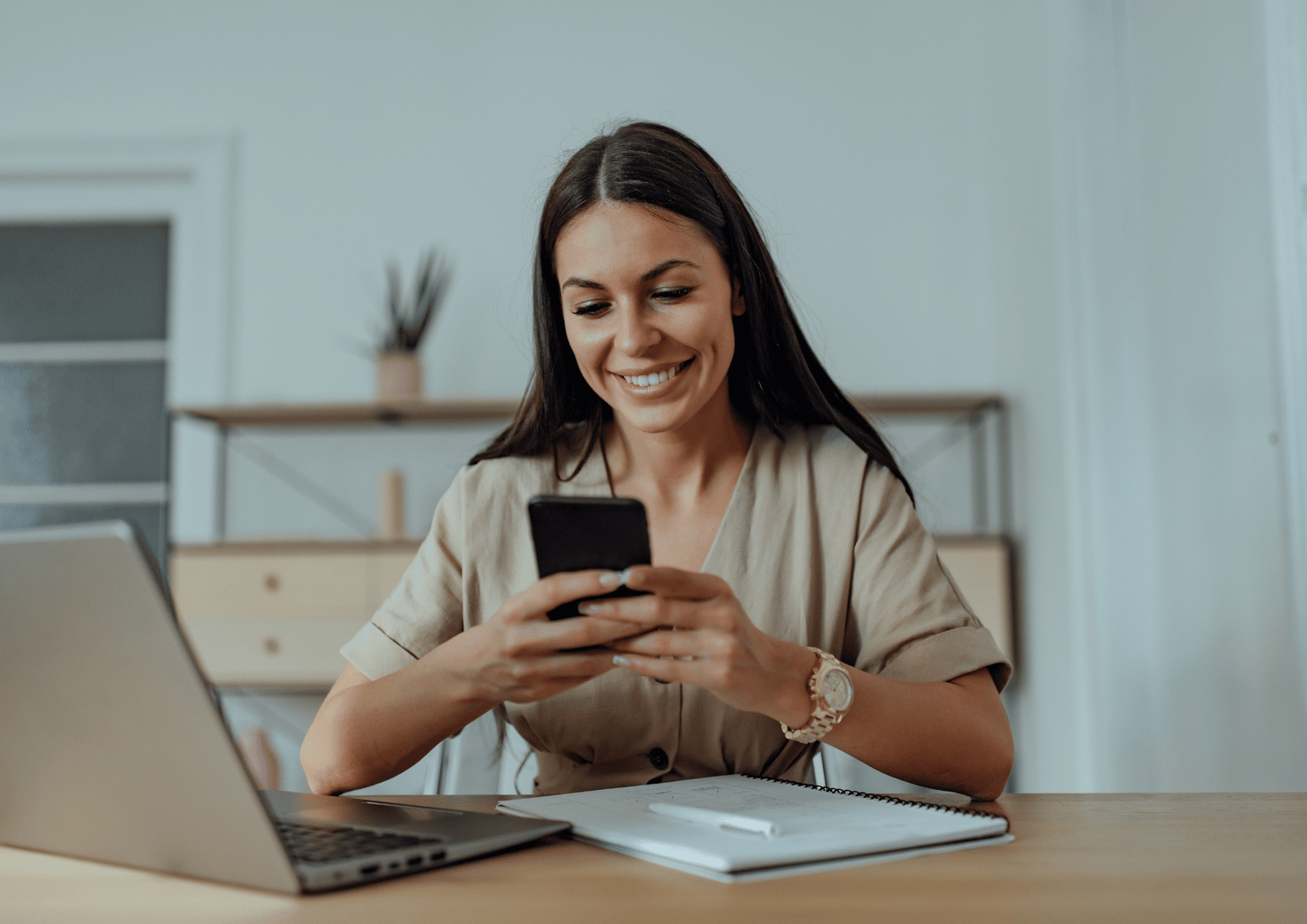 A smiling woman sitting at a desk with a laptop and notebook while checking her smartphone to send a text message.