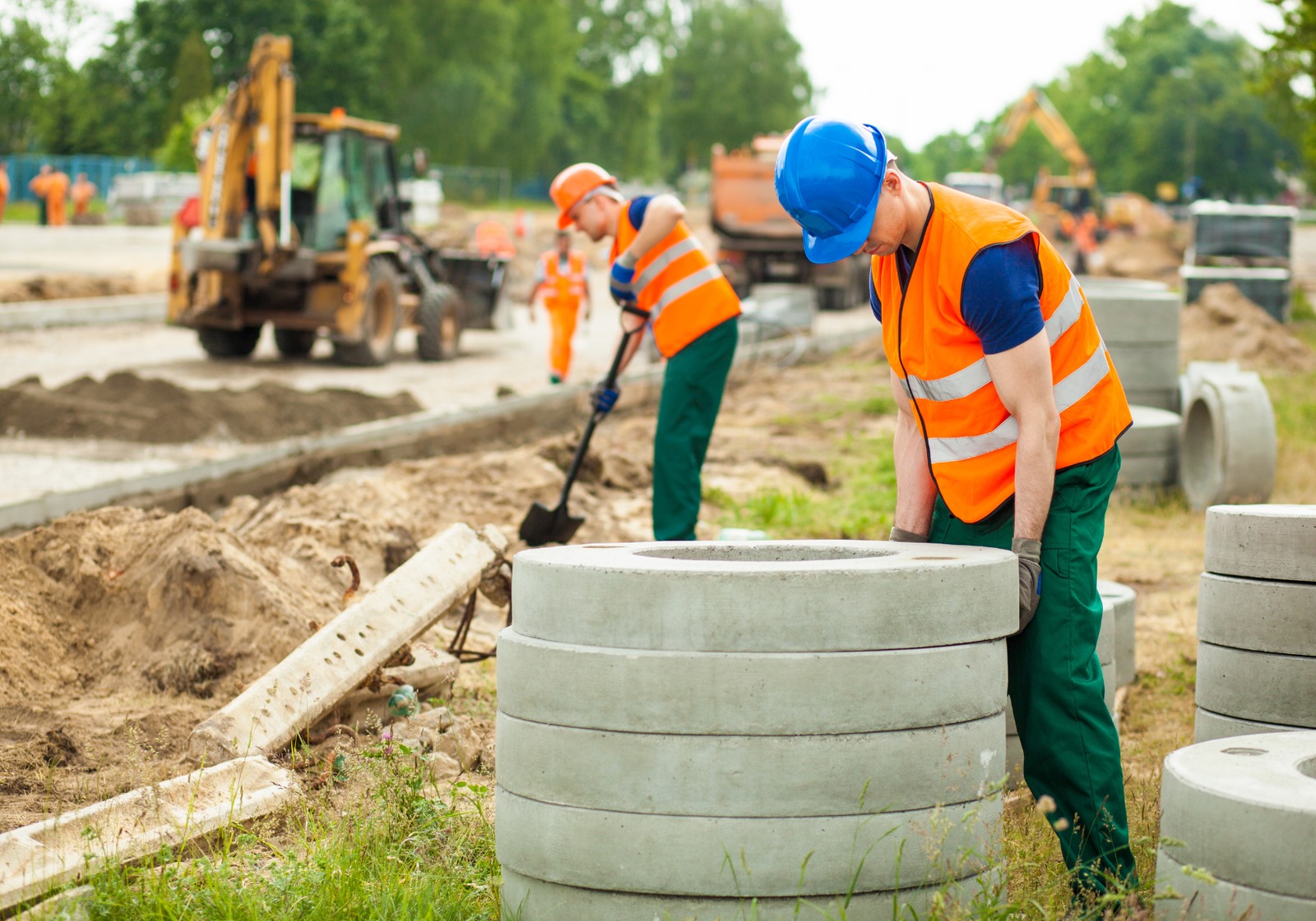 Construction workers in hardhats and safety vests lifting heavy concrete pipes at an outdoor worksite.