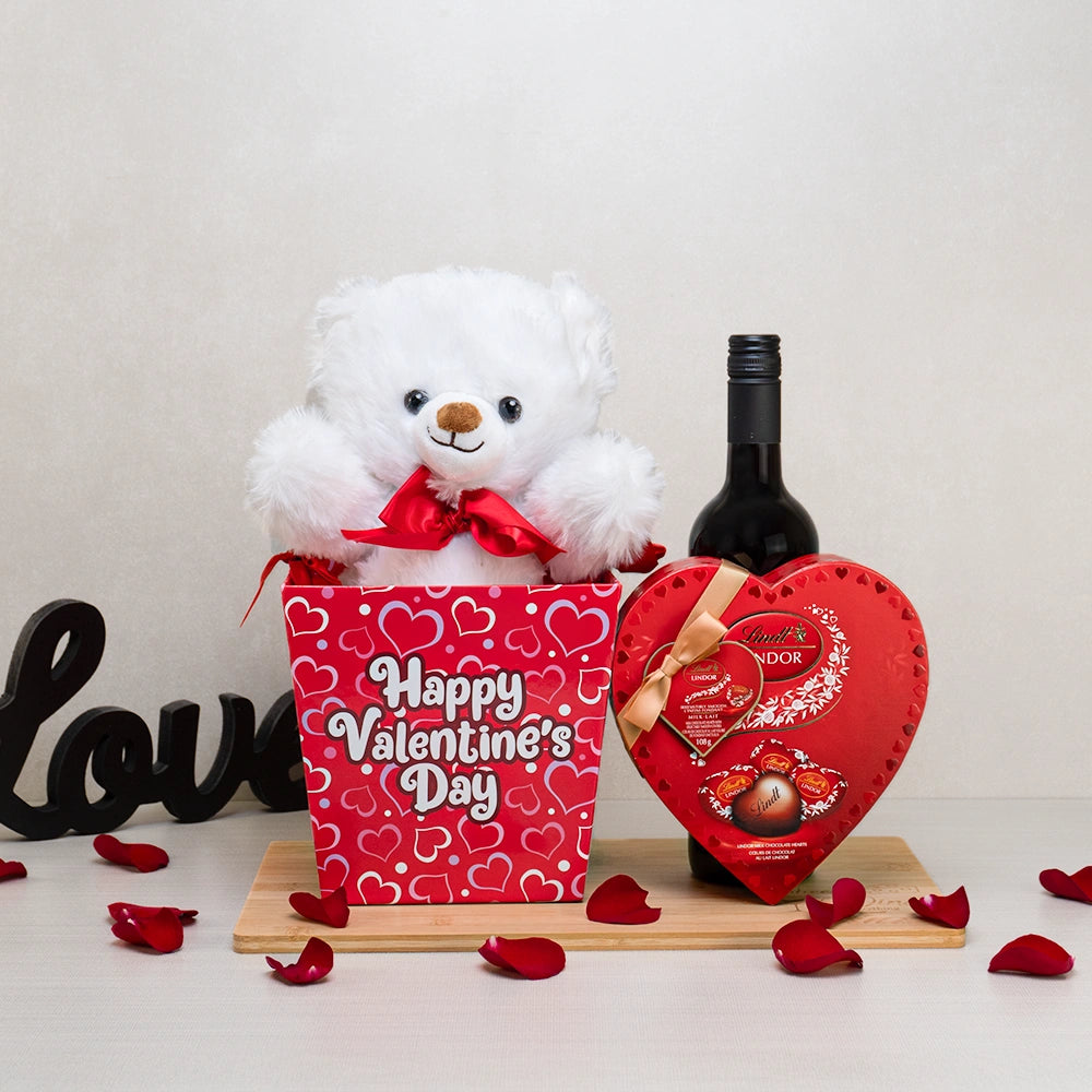 Valentines Day Gift Set with a white teddy bear in a heart-print box, red wine and a Lindt Lindor heart box on bamboo